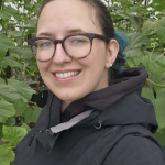 Headshot of April Norsby, woman with glasses, black jacket and background with green leaves