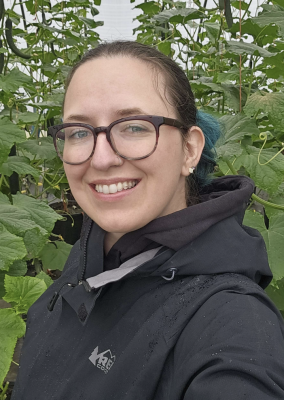 Headshot of April Norsby, woman with glasses, black jacket and background with green leaves